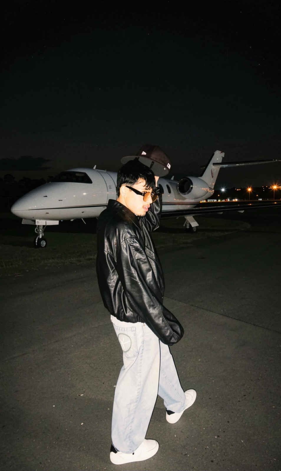 Perseo de noche en una pista de aeropuerto, con chaqueta de cuero y gorra, frente a un jet privado